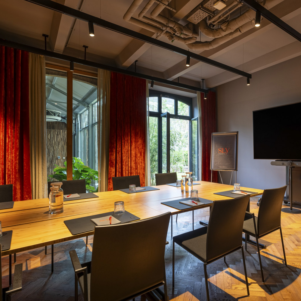 Modern conference room with a long table, chairs, a large screen, flip chart, and floor-to-ceiling windows with red curtains.