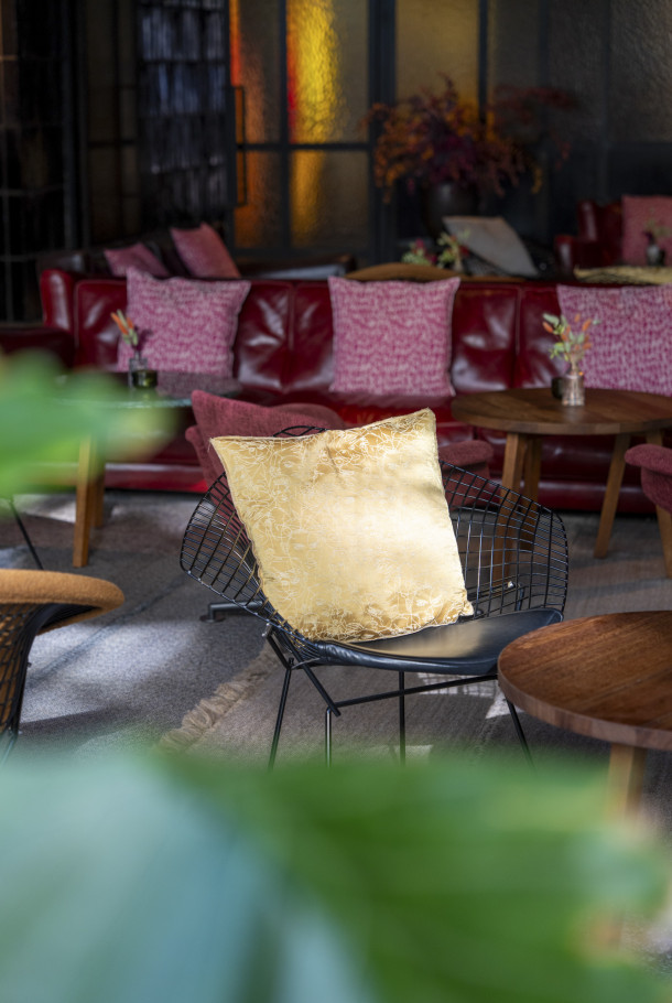 Ein gelbes Kissen auf einem schwarzen Drahtstuhl in einer gemütlichen Lounge mit roten Sofas und kleinen Holztischen im Hintergrund.