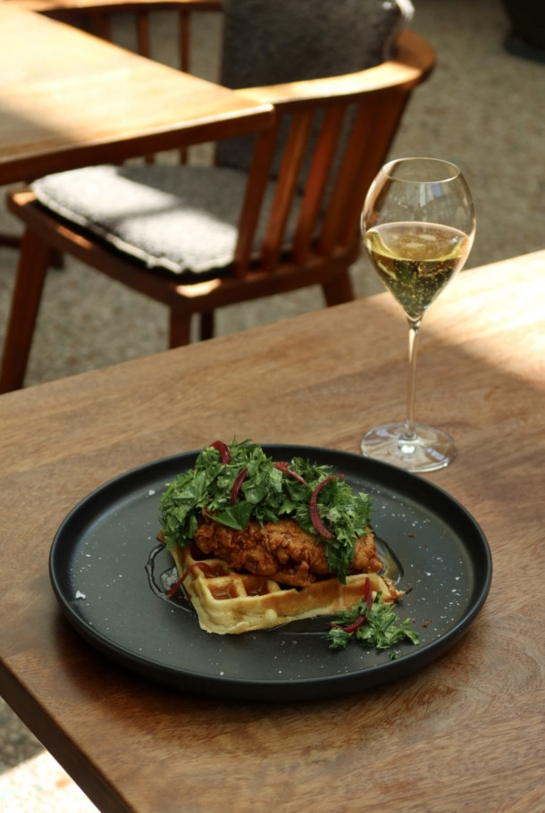 A black plate with chicken and waffles topped with greens, next to a glass of white wine on a wooden table.