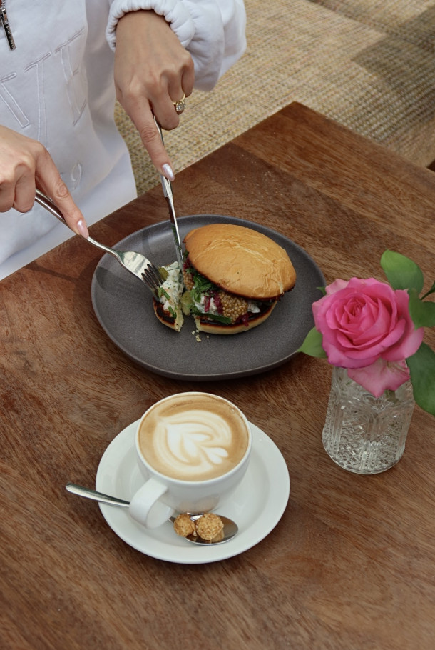 A person cuts a sandwich on a plate next to a cappuccino, chocolates, and a pink rose in a glass vase on a wooden table.