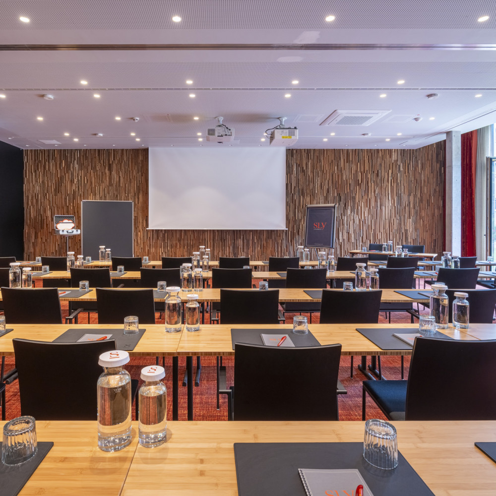 A modern conference room with rows of tables, chairs, bottled water, notebooks, and a projection screen at the front.