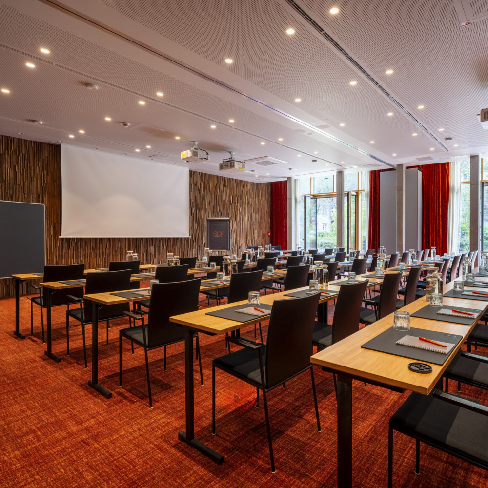 Conference room with rows of tables and chairs facing a projector screen, large windows, and red carpet flooring.