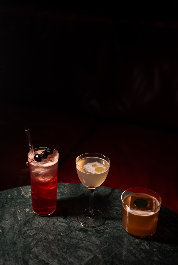 Drei Cocktails – ein roter Drink mit Kirschen, ein hellgelber Drink in einer Coupette und ein bernsteinfarbener Drink – stehen auf einem grünen Marmortisch.