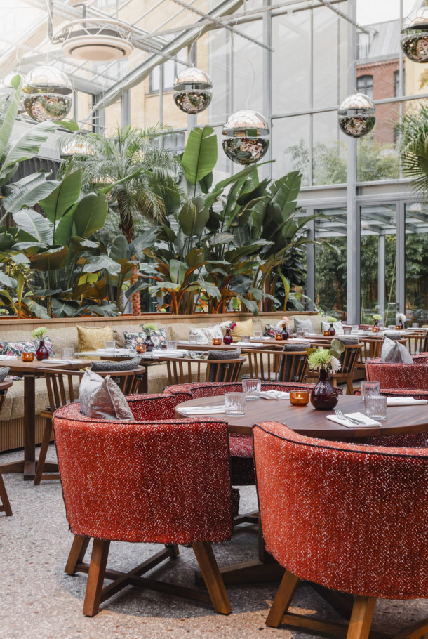 Ein stilvolles Restaurant mit roten Samtstühlen, Holztischen, üppigen grünen Pflanzen und großen Fenstern, die natürliches Licht hereinlassen.