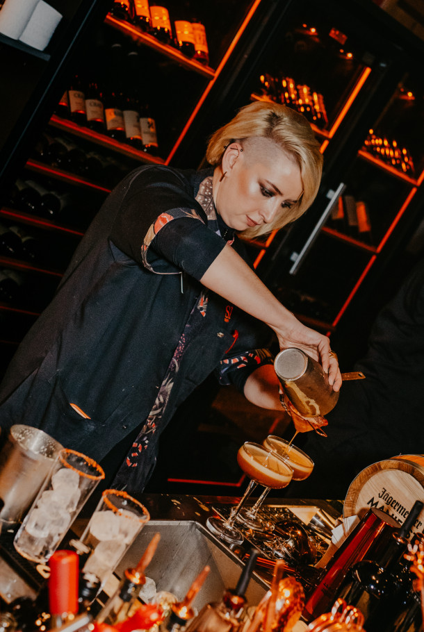 Ein Barkeeper gießt einen Cocktail in Gläser ein, im Hintergrund Flaschen und Weinregale.