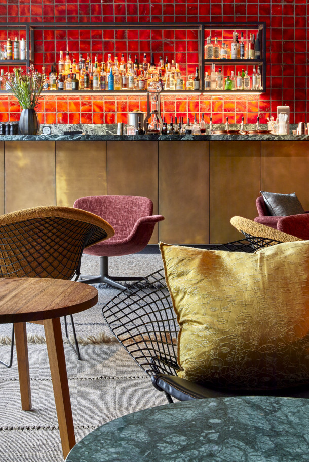 Schicke Bar mit rot gefliester Wand, Messingtheke, Flaschenregalen, modernen Stühlen und einem gelb gemusterten Kissen im Vordergrund.