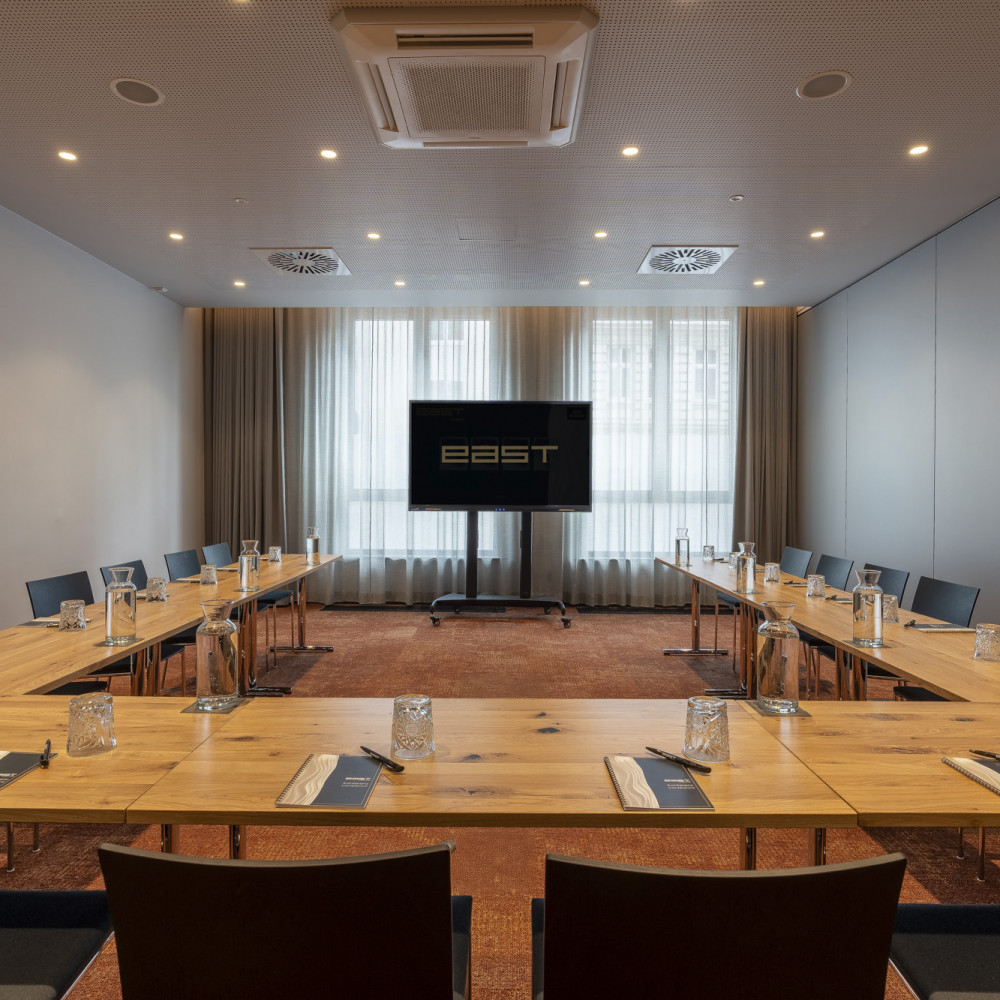 Ein moderner Konferenzraum mit U-förmigen Tischen, Stühlen, einem Fernsehbildschirm, Notizblöcken und Gläsern, beleuchtet durch Deckenleuchten und Tageslicht.