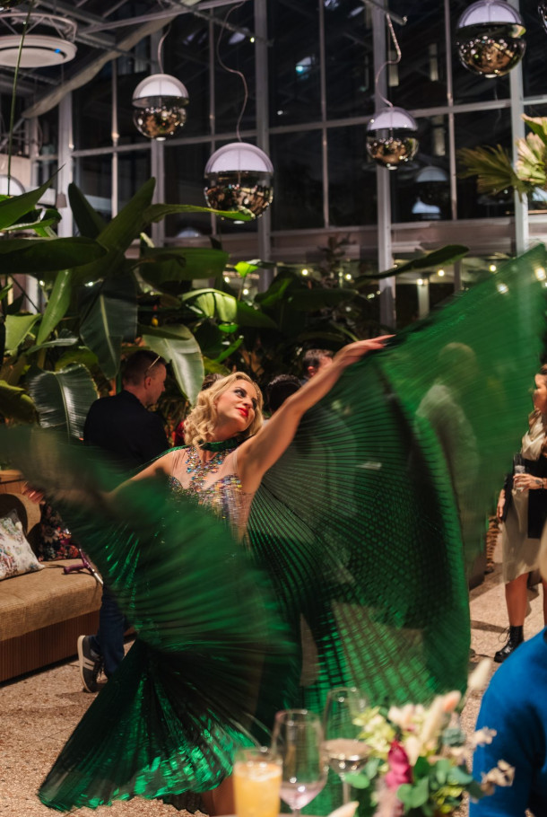 A dancer in a green costume performs indoors, surrounded by plants and hanging lights at an evening event.