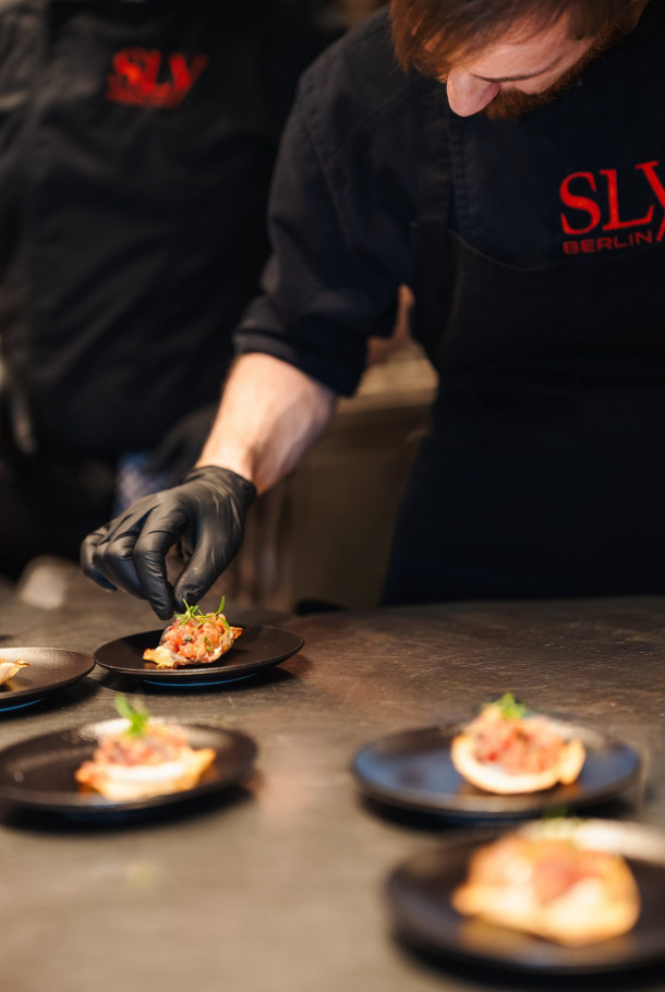 Chef in black gloves garnishes food on small black plates in a professional kitchen, wearing a black uniform with red lettering.