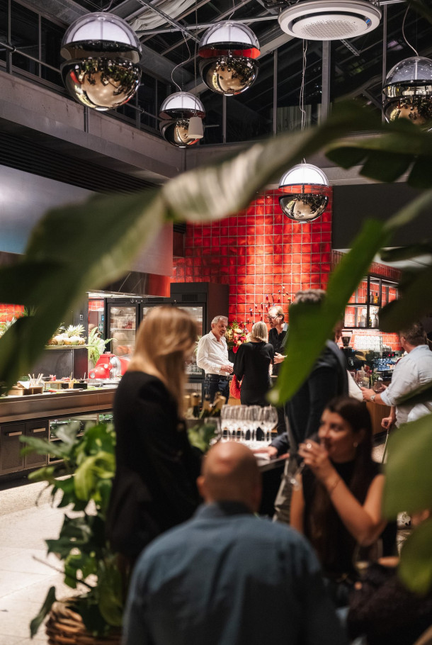People socializing in a modern, warmly lit restaurant with plants and reflective hanging lights, seen through leafy foliage.