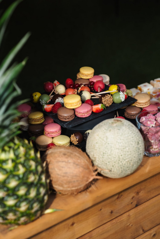 A wooden table with pineapple, coconut, melon, macarons, and chocolate-covered fruits on display.
