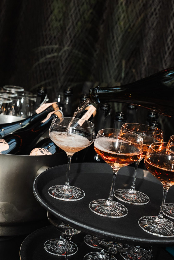A bottle pours sparkling wine into coupe glasses on a tray, with more bottles chilling in an ice bucket nearby.