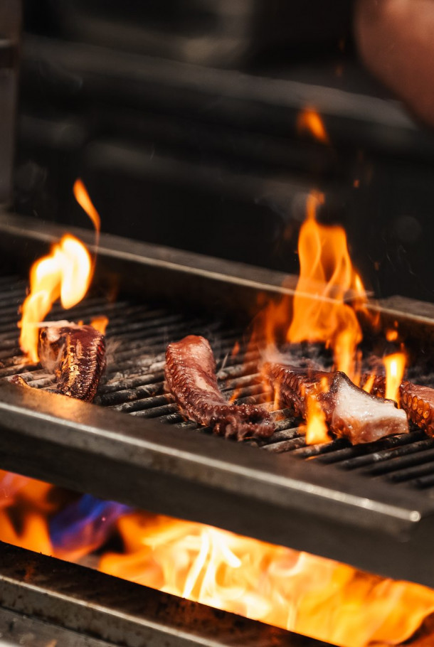 Flames rise as various pieces of meat cook on a grill, with a persons hand visible in the background.