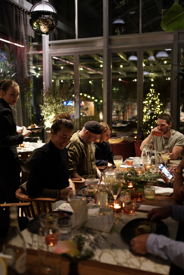 Eine Gruppe von Menschen genießt gemeinsam das Abendessen in einem warm beleuchteten Restaurant, während ein Kellner ihren Tisch bedient und im Hintergrund festliche Lichter leuchten.