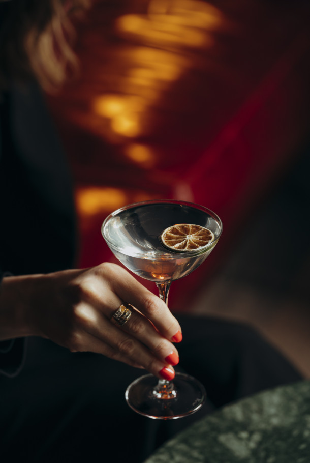 Eine Hand mit roten Nägeln hält vor einem warmen, sanft beleuchteten Hintergrund ein Martini-Glas mit einer Garnitur aus getrockneten Zitrusscheiben.