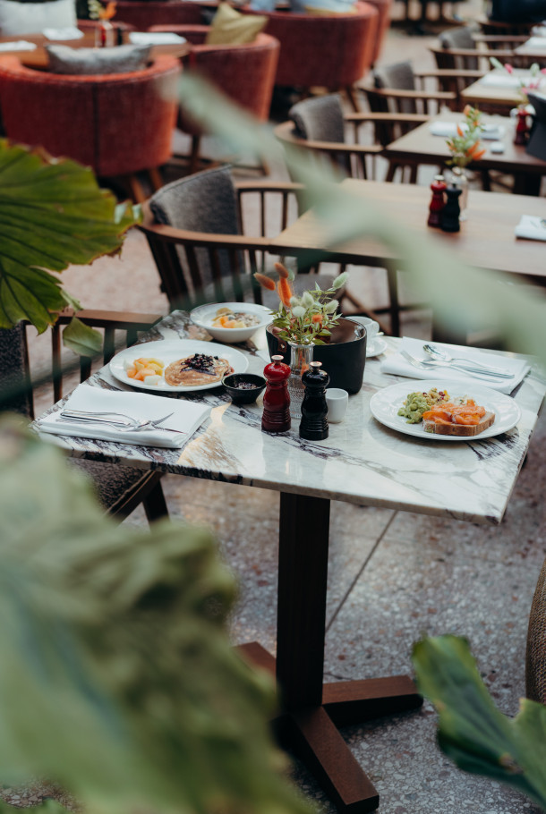 Ein für zwei Personen gedeckter Marmortisch mit Tellern voller Essen, Getränken und Gewürzen in einem Freiluftrestaurant, umgeben von Pflanzen und Stühlen.