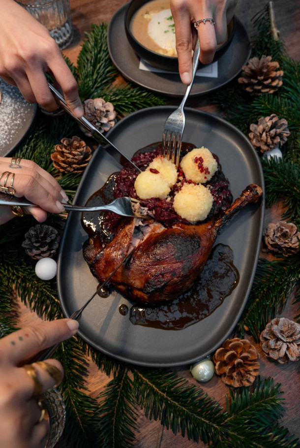 Three hands cut into roasted duck served with dumplings and purple cabbage, garnished with pine cones and fir branches on a festive table.