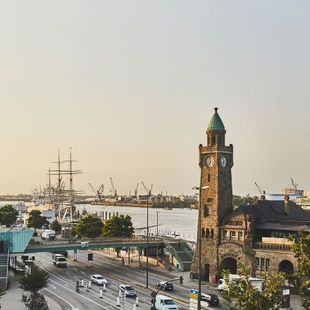 Blick auf einen Hafen mit einem Uhrenturm, Schiffen, Kränen, Autos und Menschen in der Nähe der Uferpromenade an einem klaren Tag.
