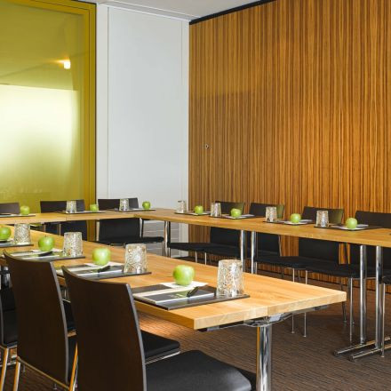 A modern conference room with U-shaped tables, black chairs, green apples, glasses, and notepads neatly set at each seat.