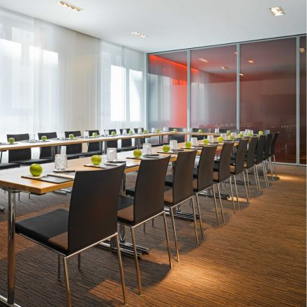 A modern conference room with two long tables, black chairs, notepads, glasses, and green apples neatly arranged at each seat.