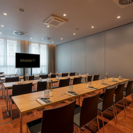 A modern conference room with rows of tables and chairs, notepads, glasses, and a large screen displaying the word EAST.