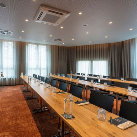 Modern conference room with long wooden tables, black chairs, notepads, pens, glasses, and water pitchers, with large curtained windows.