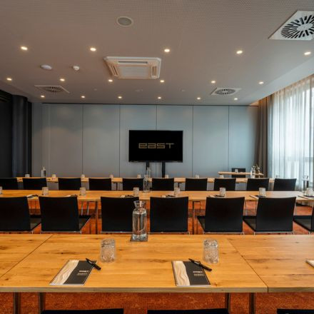 Modern conference room with rows of tables and chairs facing a screen, notepads and water bottles on each table, and large windows to the right.