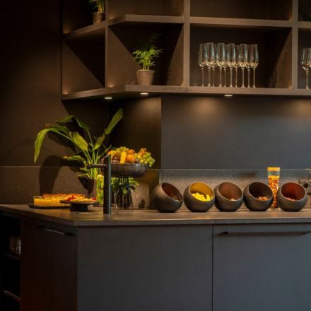 Modern kitchen counter with fruit bowls, leafy plant, glasses on shelves, and warm under-cabinet lighting.