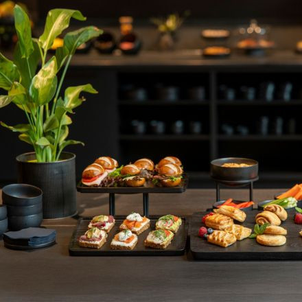 A variety of appetizers, sandwiches, and pastries are displayed on trays beside a potted plant on a dark wooden table.