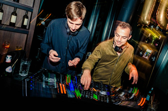 Zwei DJs treten bei einer Party auf, im Hintergrund sind goldene Luftballons und Regale voller Weinflaschen zu sehen.