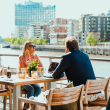 Ein Paar speist an einem sonnigen Tag im Freien in einem Restaurant am Flussufer mit modernen Gebäuden und Wasser im Hintergrund.