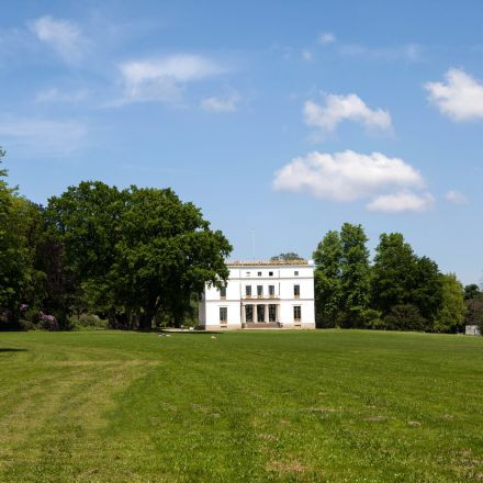 Ein weißes, zweistöckiges Gebäude steht inmitten einer großen grünen Wiese mit Bäumen unter einem blauen Himmel mit vereinzelten Wolken.