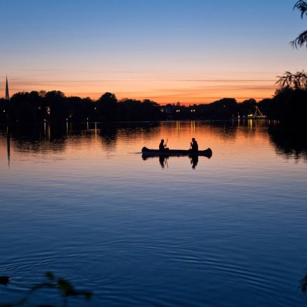 Zwei Personen fahren bei Sonnenuntergang Kanu auf einem ruhigen See, im Wasser spiegeln sich Bäume und ein bunter Himmel.