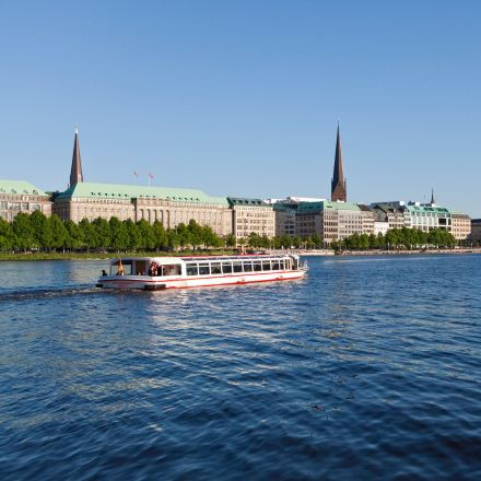 Ein weißes Boot fährt auf einem ruhigen Fluss, im Hintergrund sind unter einem klaren blauen Himmel Stadtgebäude und Kirchtürme zu sehen.