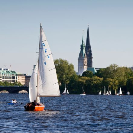 Ein rotes Segelboot auf einem Fluss mit anderen Segelbooten und einer Stadtlandschaft, darunter eine hohe Kirche, im Hintergrund.