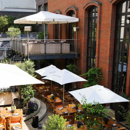 Outdoor restaurant patio with wooden tables and white umbrellas next to a brick building and large windows, with greenery throughout.
