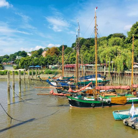 Bunte Segelboote liegen an einem Flussufer vor Anker, im Hintergrund sind üppig grüne Bäume und ein blauer Himmel zu sehen.
