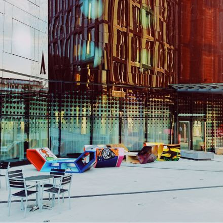 Bunte, geometrische Bänke und leere Stühle stehen auf einem modernen Platz mit reflektierenden Glasgebäuden im Hintergrund.