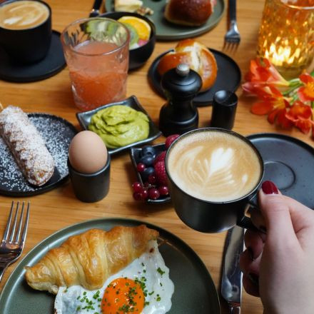 A breakfast spread with croissant, fried egg, berries, avocado, soft-boiled egg, coffee, juice, and pastries on a wooden table.