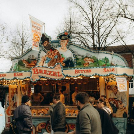 Auf einem Markt im Freien gehen Menschen an einem festlichen Brezelstand vorbei, der mit traditionellen deutschen Kunstwerken und hellen Lichtern dekoriert ist.