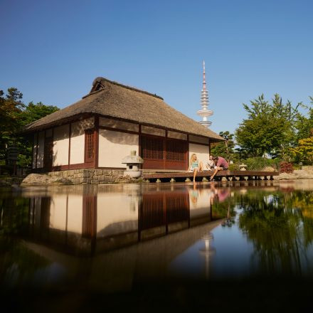 Ein traditionelles japanisches Teehaus mit Strohdach, das sich in einem Teich spiegelt, mit einer Pagode und Bäumen im Hintergrund unter einem klaren Himmel.