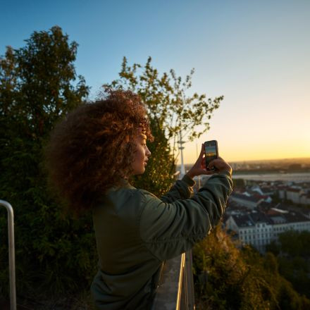 Eine Person mit lockigem Haar macht bei Sonnenuntergang mit einem Smartphone ein Selfie und blickt von einem hohen Aussichtspunkt auf eine Stadtlandschaft.