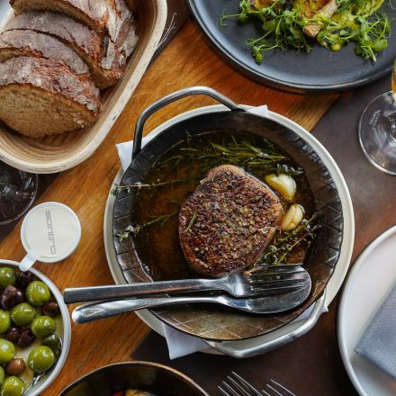 A cooked steak with herbs and garlic in a pan, served with bread, olives, and salad on a wooden table.