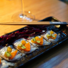 Sliced fish topped with orange pieces and greens, served on a black plate with chopsticks and red cabbage on a wooden table.