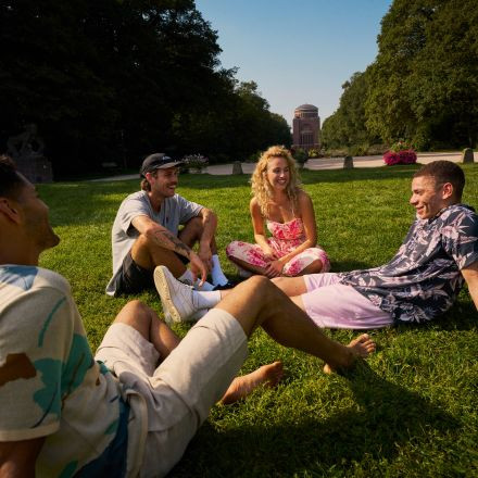 An einem sonnigen Tag sitzen vier Freunde lächelnd und unterhalten sich auf dem Gras in einem Park. Im Hintergrund sind Bäume und ein Gebäude zu sehen.