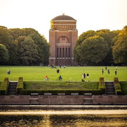 Menschen gehen und fahren mit dem Fahrrad auf einer Rasenfläche vor einem großen Backsteingebäude, das von Bäumen umgeben ist, im Vordergrund ist Wasser zu sehen.