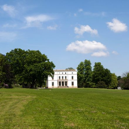 In der Ferne steht ein weißes Herrenhaus, umgeben von grünem Gras, Bäumen und einem blauen Himmel mit vereinzelten Wolken.
