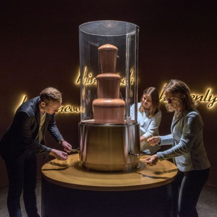 Drei Personen versammeln sich um einen großen Schokoladenbrunnen und tunken Leckereien ein, während im Hintergrund Neonschilder leuchten.