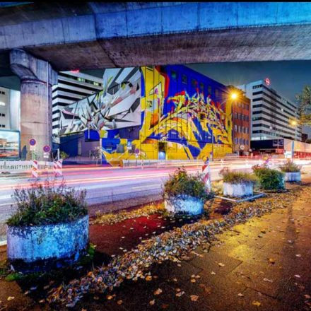 Ein lebendiges Wandgemälde mit abstrakten Formen schmückt nachts ein Gebäude an einer belebten Stadtstraße mit Lichtspuren vorbeifahrender Autos.