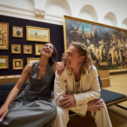 Zwei Menschen sitzen lachend auf einer Bank in einer Kunstgalerie, während an den Wänden hinter ihnen verschiedene gerahmte Gemälde hängen.